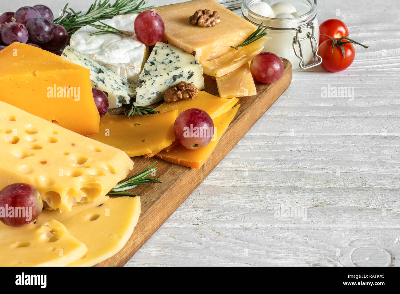 different types of cheeses on wooden cutting board. Camembert cheese, cheddar, hard cheese slices, soft cheese, walnuts, grapes, tomatoes and rosemary Stock Photo