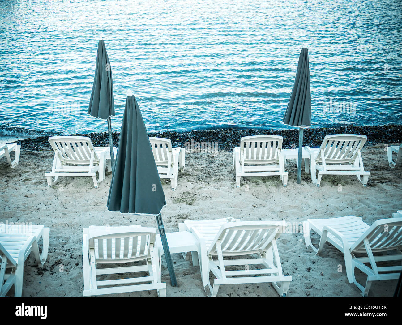 Off season on the sand beach with no people. Closed sun umbrellas, parasol, white plastic chairs and couches on seaside with sea grass Stock Photo