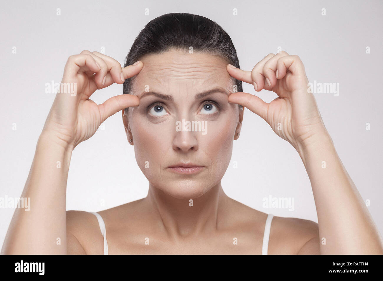 Portrait of pretty, charming, attractive middle aged woman with perfect face skin checking wrinkles on her forehead, holding fingers on brow, isolated Stock Photo
