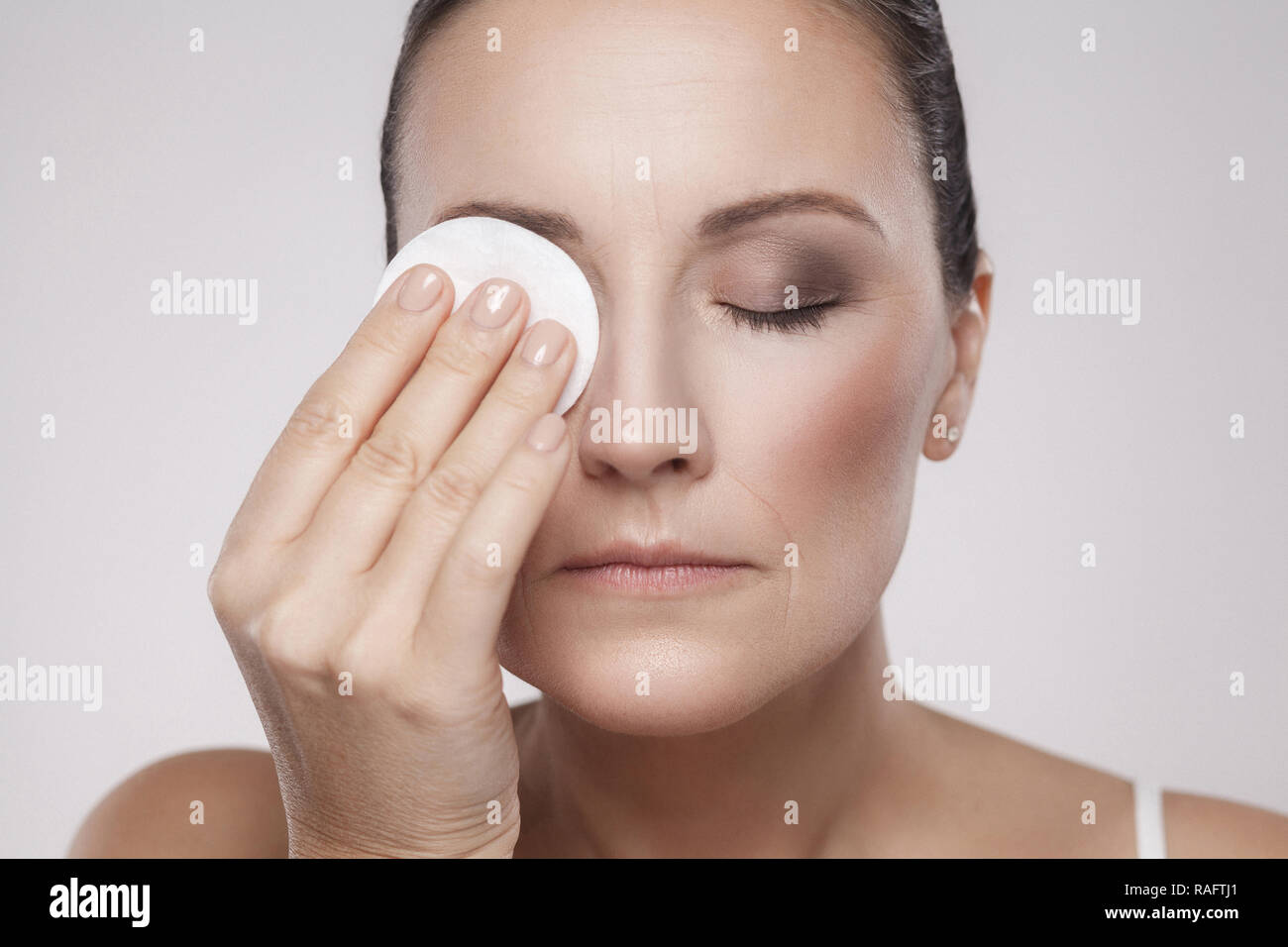 Excited cheerful confident lovely beautiful mature woman is using cotton pad with micellar water for removing make up from eye, isolated on grey backg Stock Photo