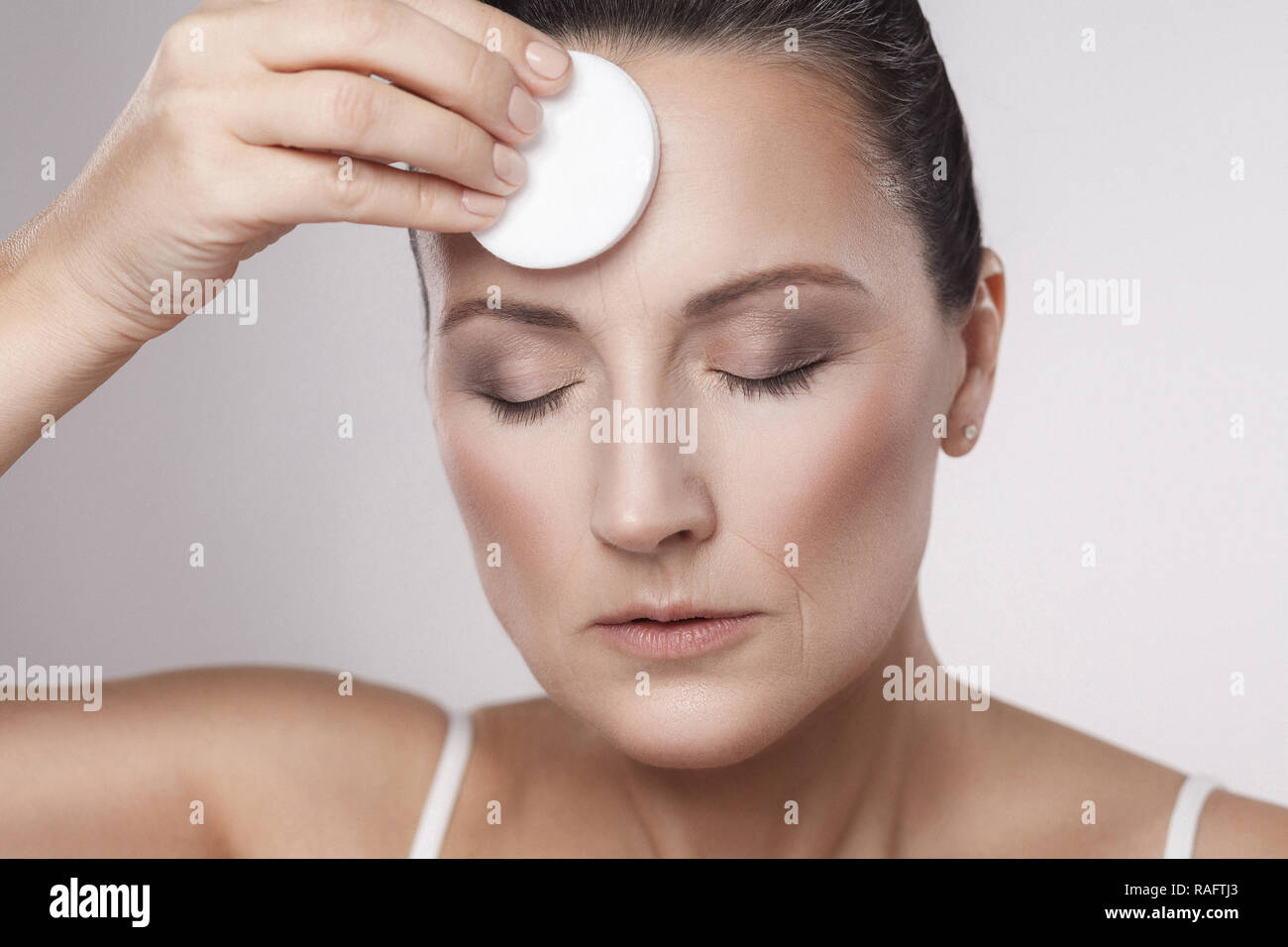 Excited cheerful confident lovely beautiful mature woman is using cotton pad with micellar water for removing make up from face, isolated on grey back Stock Photo