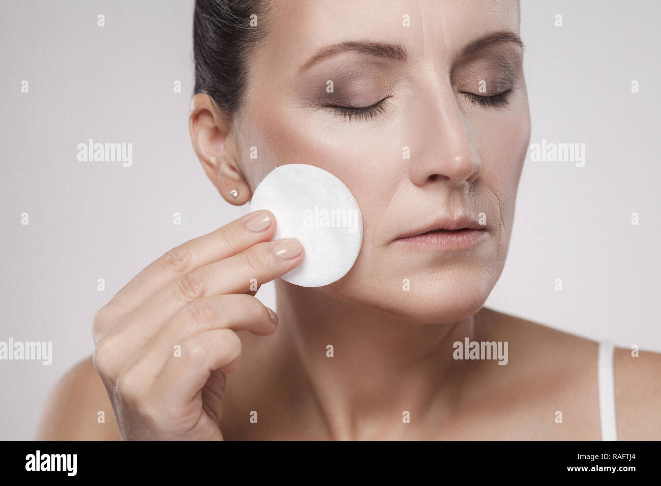 Excited cheerful confident lovely beautiful mature woman is using cotton pad with micellar water for removing make up from face, isolated on grey back Stock Photo