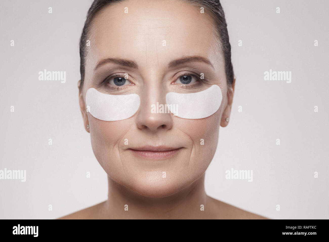Closeup portrait of beautiful middle aged woman with perfect skin using white hydrogel patches with lifting anti-wrinkle collagen effect, gray backgro Stock Photo