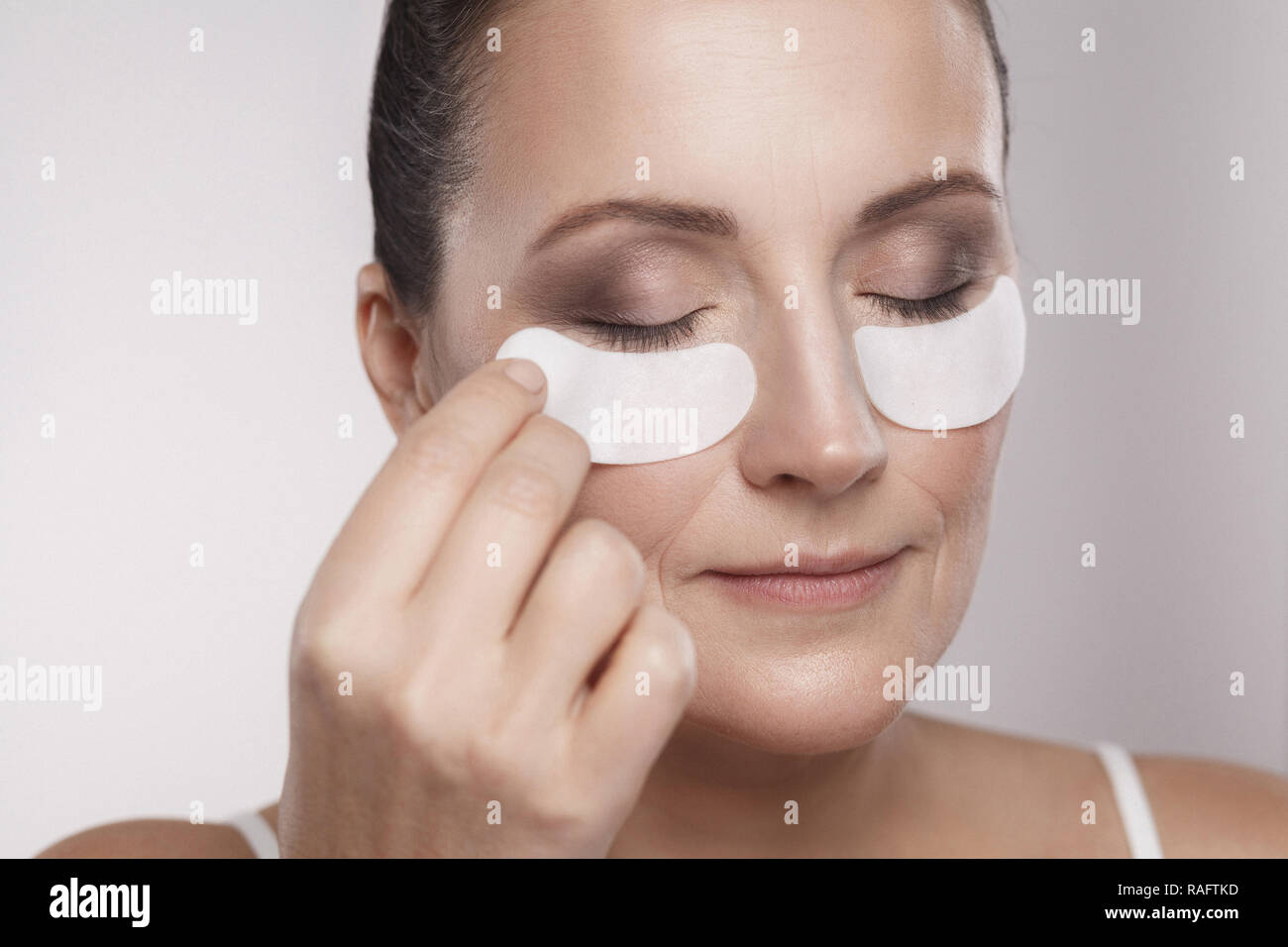 Closeup portrait of beautiful middle aged woman with perfect skin using white hydrogel patches with lifting anti-wrinkle collagen effect, gray backgro Stock Photo