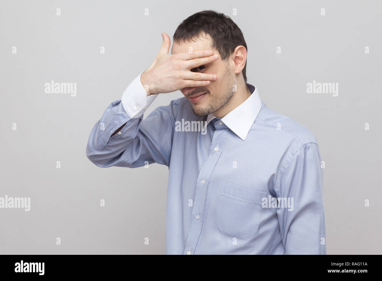 Portrait of funny shy handsome bristle businessman in classic blue shirt standing, covering his eyes and looking between fingers at camera. indoor stu Stock Photo