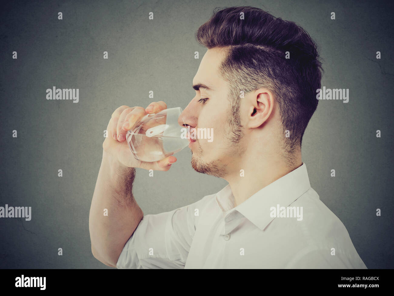Side view of modern man in white shirt drinking water from glass on gray background Stock Photo