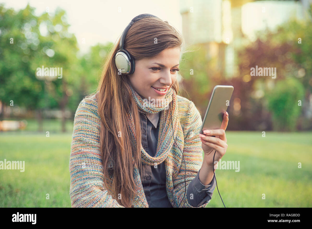 Young casual woman in headphones using phone while sitting in park and listening to music Stock Photo