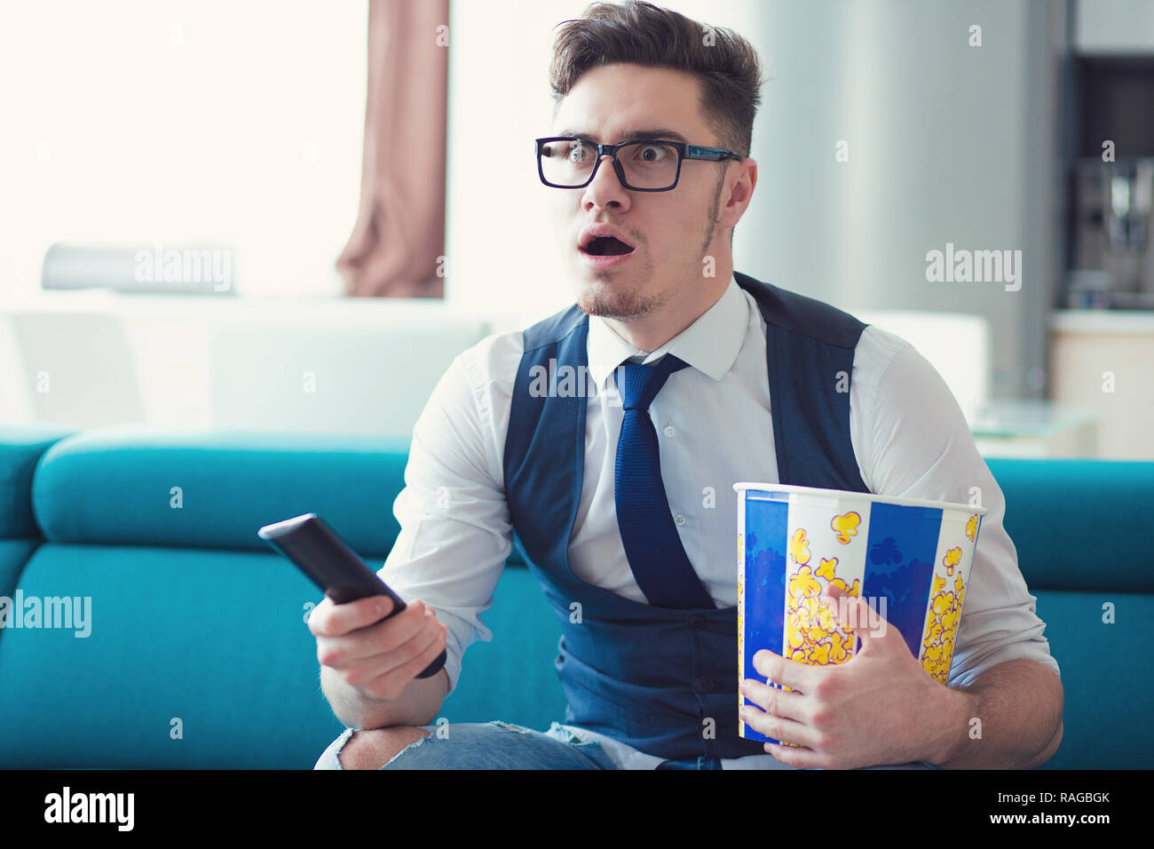 Portrait of an elegant man sitting on a couch, watching TV, holding remote and popcorn box, surprised at what he sees, isolated indoors flat backgroun Stock Photo