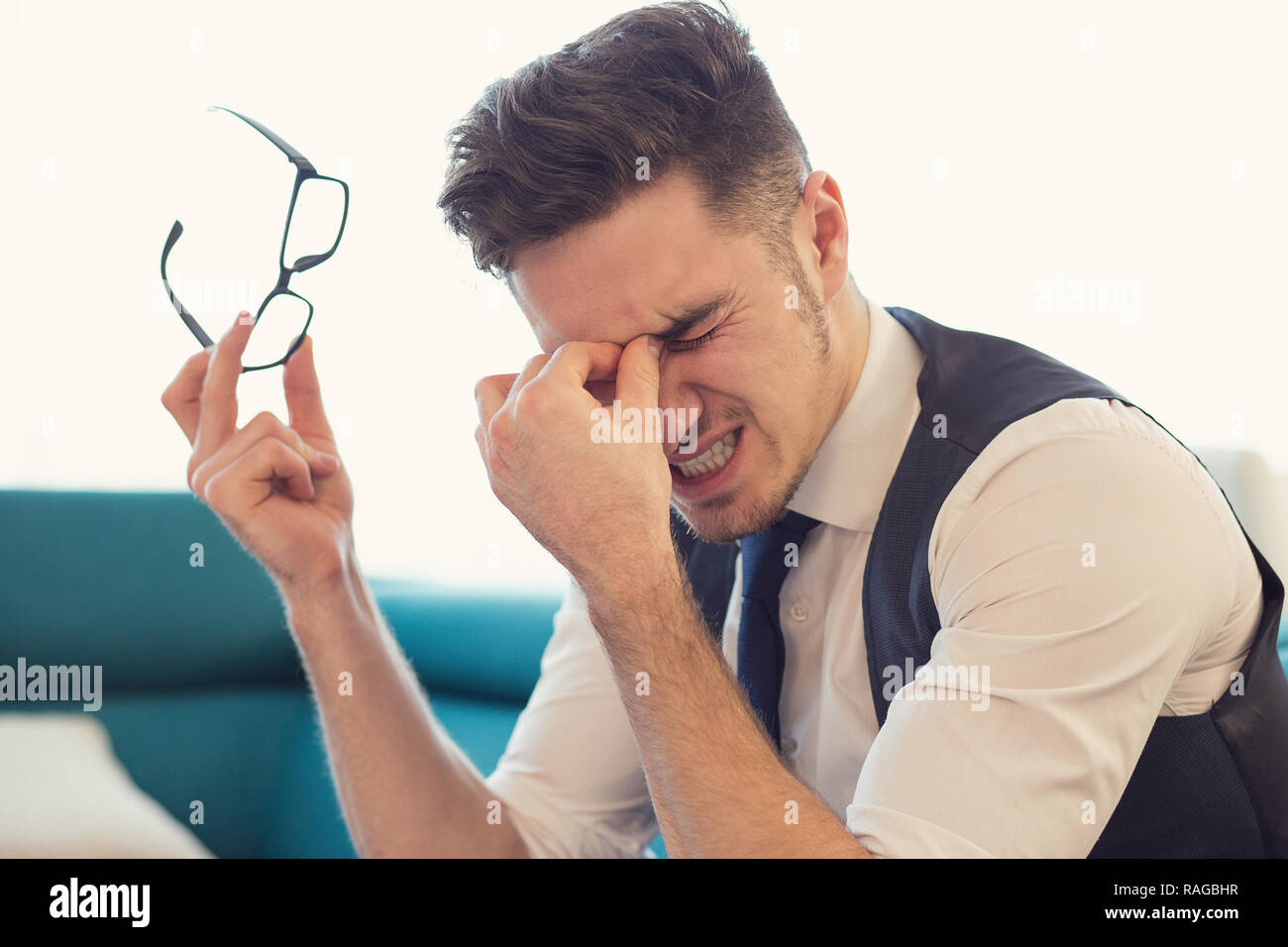 Young man rubbing nose bridge while holding eyeglasses and suffering with eyes problems sitting on couch Stock Photo