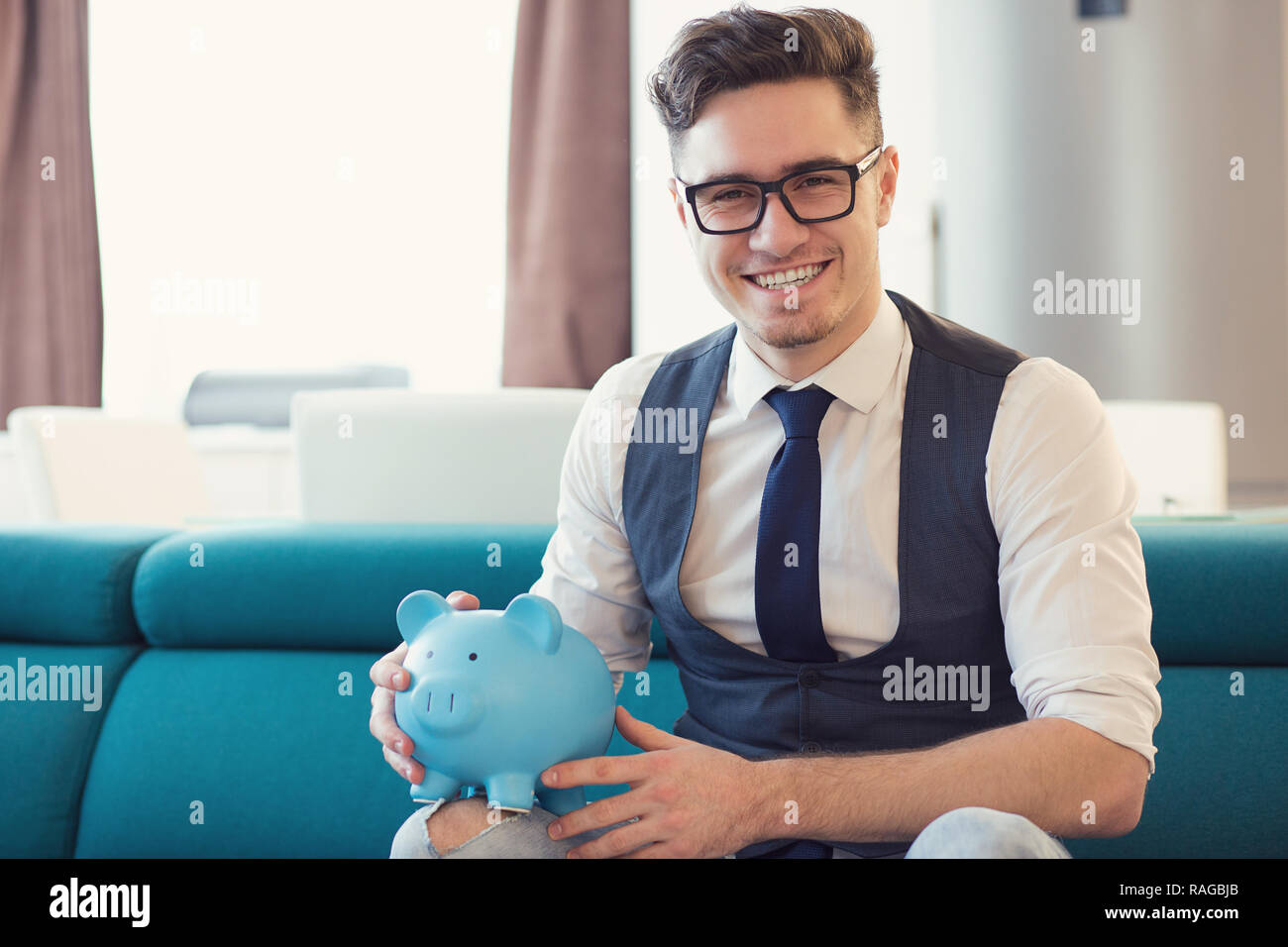 Happy formal man in glasses holding blue piggy bank sitting on sofa in new modern apartment smiling at camera Stock Photo