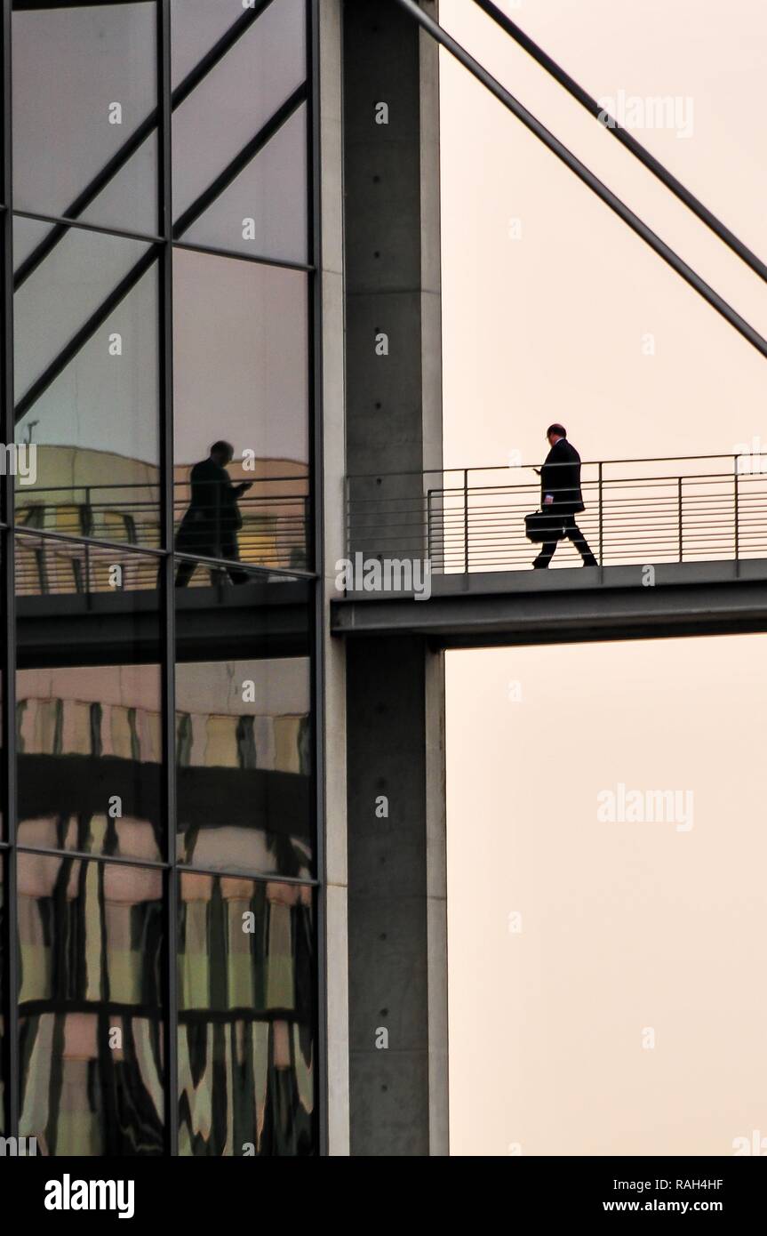 Businessman walking across bridge towards business building, nobodz, reflection in glass facade, holding a mobile phone, business and executive Stock Photo