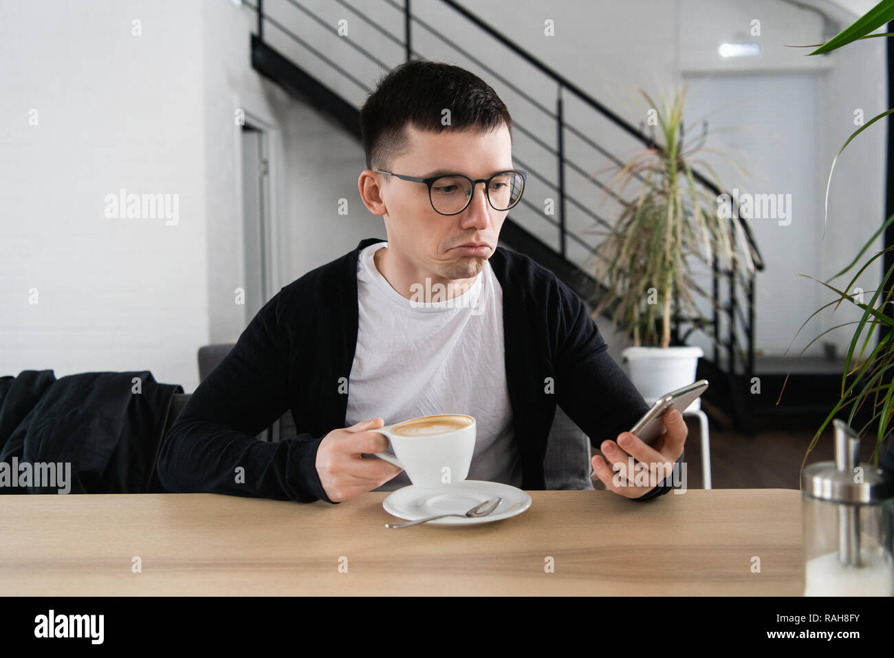 Worried young man reading a message with bad notification on modern smartphone sitting in cafe. Frustrated and upset hipster guy watching shocking video in blog on cellular Stock Photo