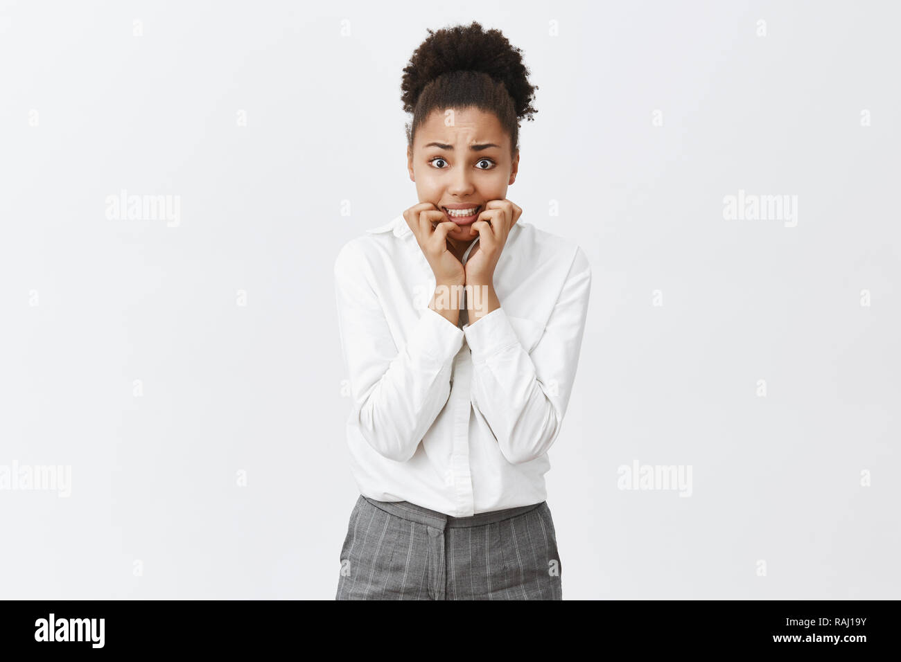 Girl stuck in elevator being scared to death, standing over gray background, biting fingernails from fear and anxiety, staring intensely at camera, waiting for rescue, wearing office pants and shirt Stock Photo