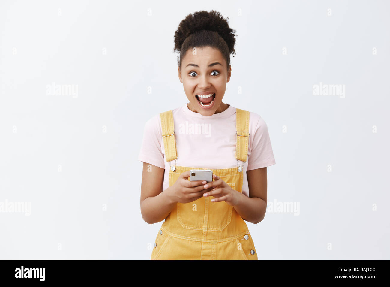 Womans creaming from emotions, receiving invitation to famous party. Portrait of surprised and amazed good-looking dark-skinned girl in yellow overalls, yelling at camera and holding smartphone Stock Photo