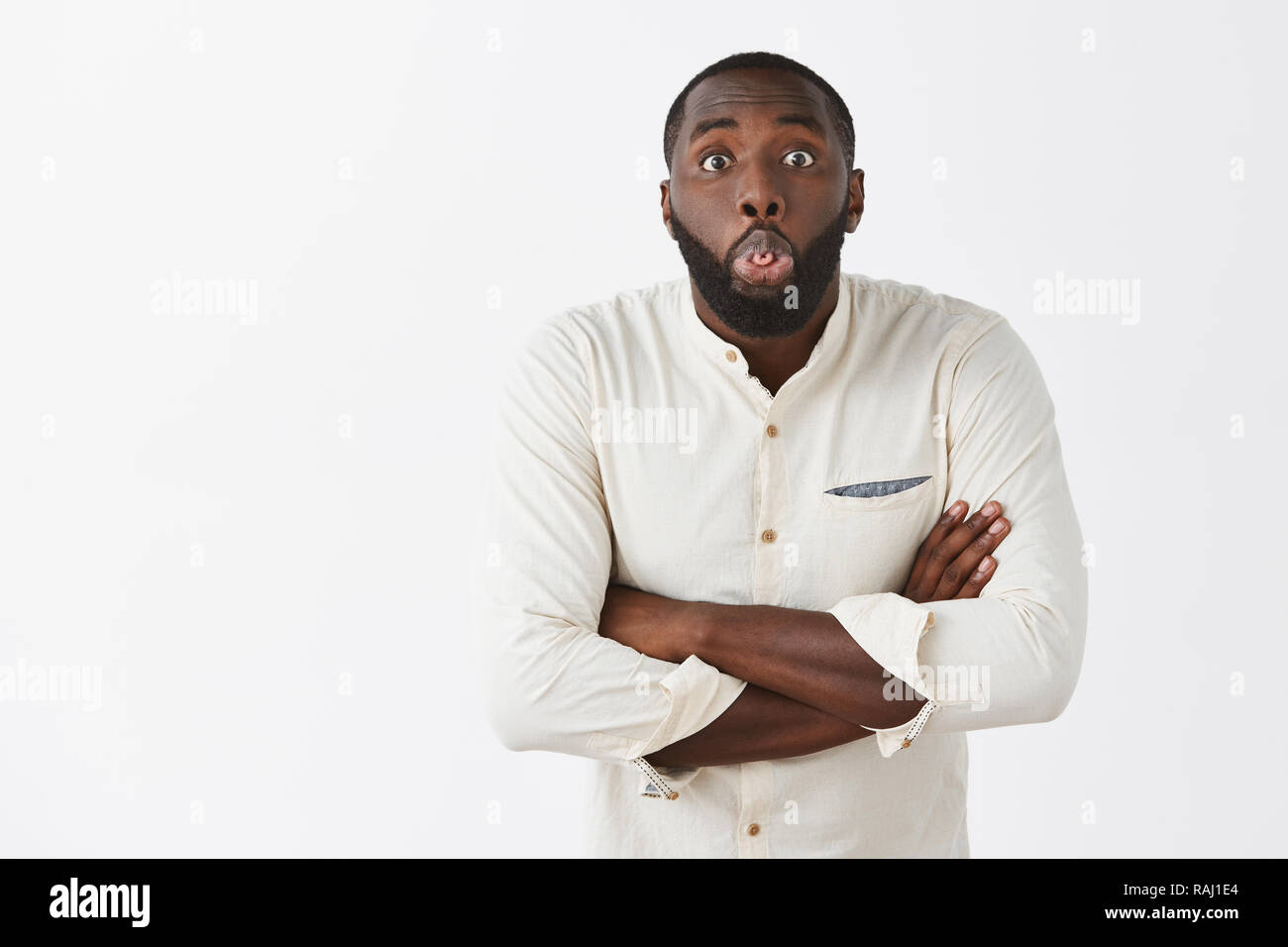 Indoor shot of cute and funny African American male in white trendy shirt mocking girlfriend, rolling and showing tongue, holding hands crossed, being childish and joyful over gray background Stock Photo