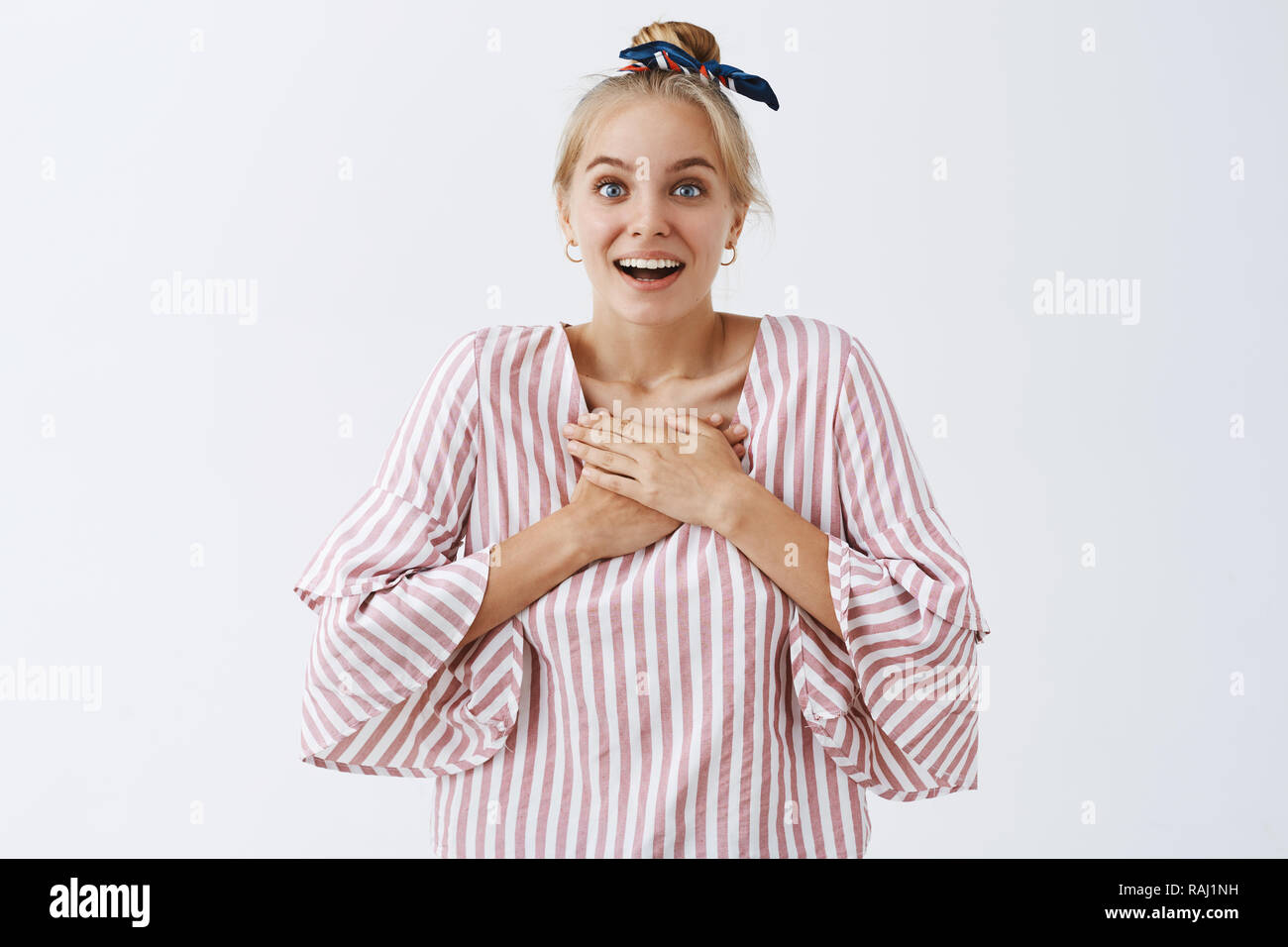 You make heart beat faster. Portrait of charmed and amazed cute modern woman in striped blouse and headband, smiling broadly, holding palms on chest and smiling, gasping, losing speech from surprise Stock Photo