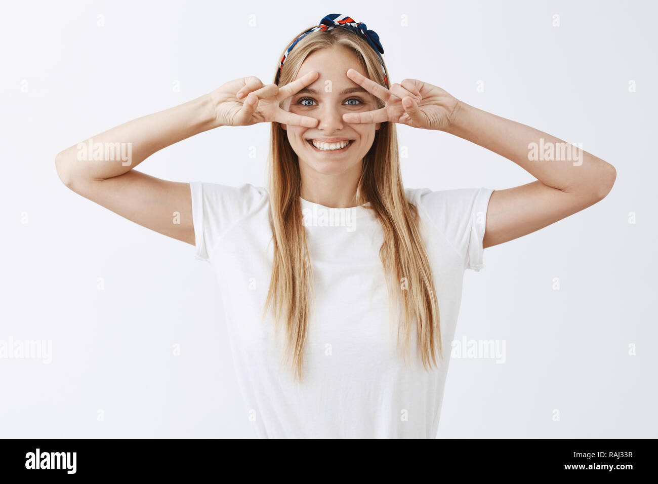 Smile never been so cute. of charming and happy carefree european girl with  blond hair, showing victory or peace signs over eyes and grinning, being in  playful and joyful mood over grey
