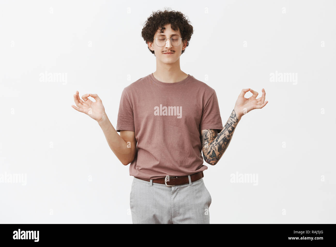 Charming curly-haired guy with funny moustache standing in lotus pose trying find peace and comfort closing eyes and smiling with calm and relaxed expression meditating over gray background Stock Photo