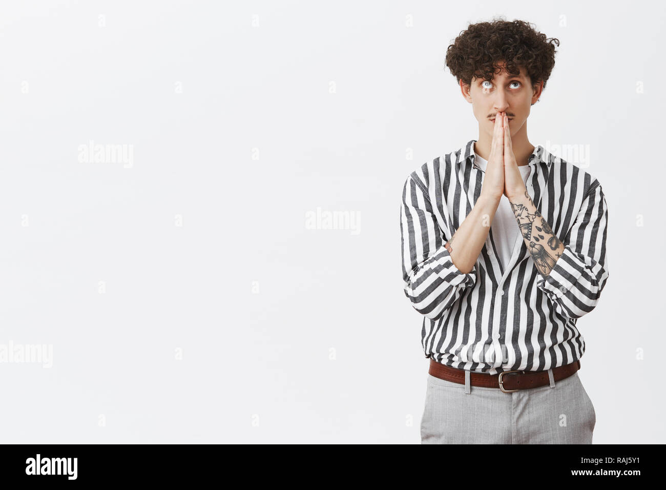 Indoor shot of cute young and stylish jewish guy with dark curly hair moustache and tatoos in striped shirt and pants holding hands in pray looking up while making wish having faith in god Stock Photo