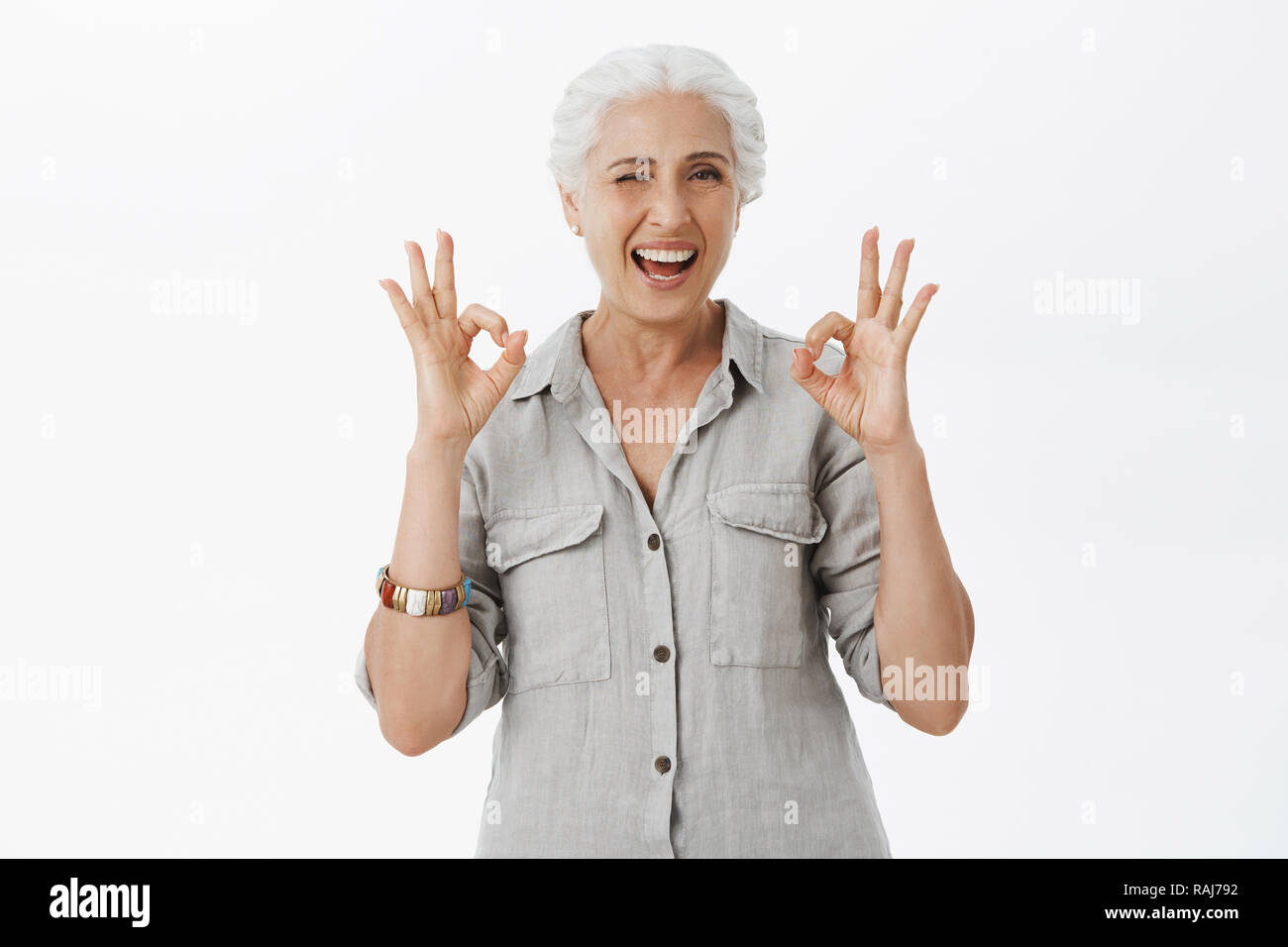 Waist-up shot of charismatic happy and cute awesome granny winking joyfully smiling and showing okay gestures confirming she dealt with everything and we not have worry over gray background Stock Photo