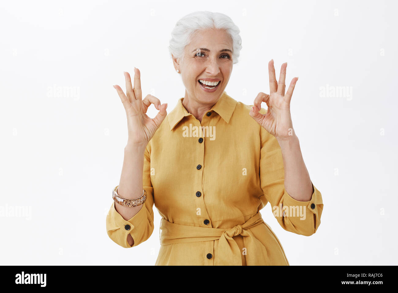 granny posing Granny has excellent health. Portrait of joyful active and emotive senior  woman with white hair smiling joyfully and laughing showing okay or perfect  gesture posing in trendy yellow coat Stock Photo -