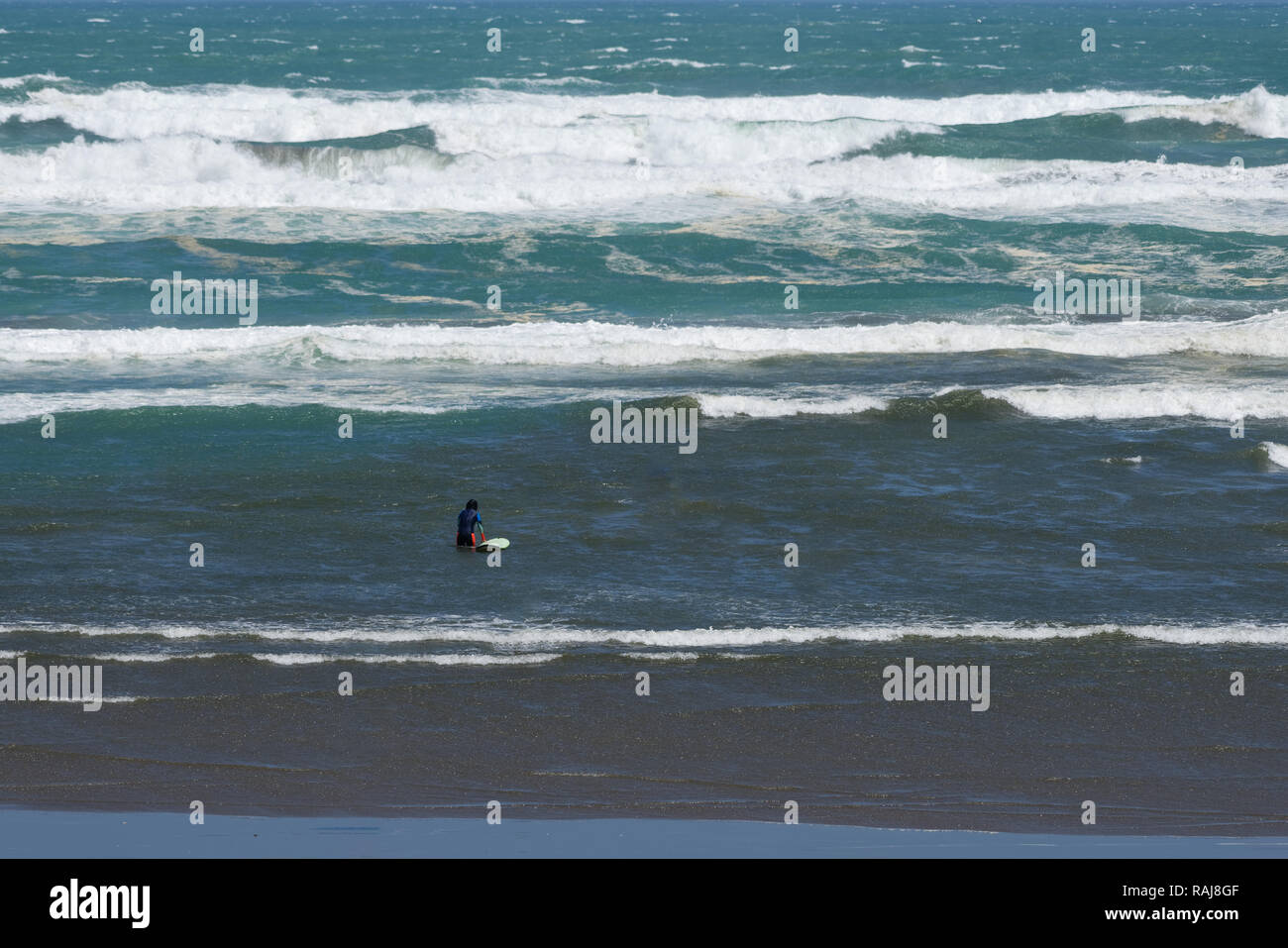 A lone surfer walking into the rough surf on a windy day Stock Photo
