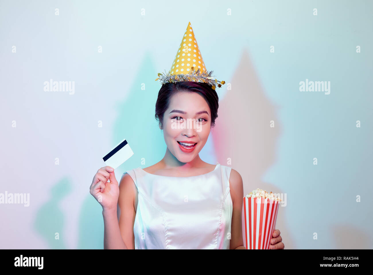 Portrait of young overjoyed attractive woman in white dress watching movie film, holding bucket of popcorn and credit card isolated on white backgroun Stock Photo