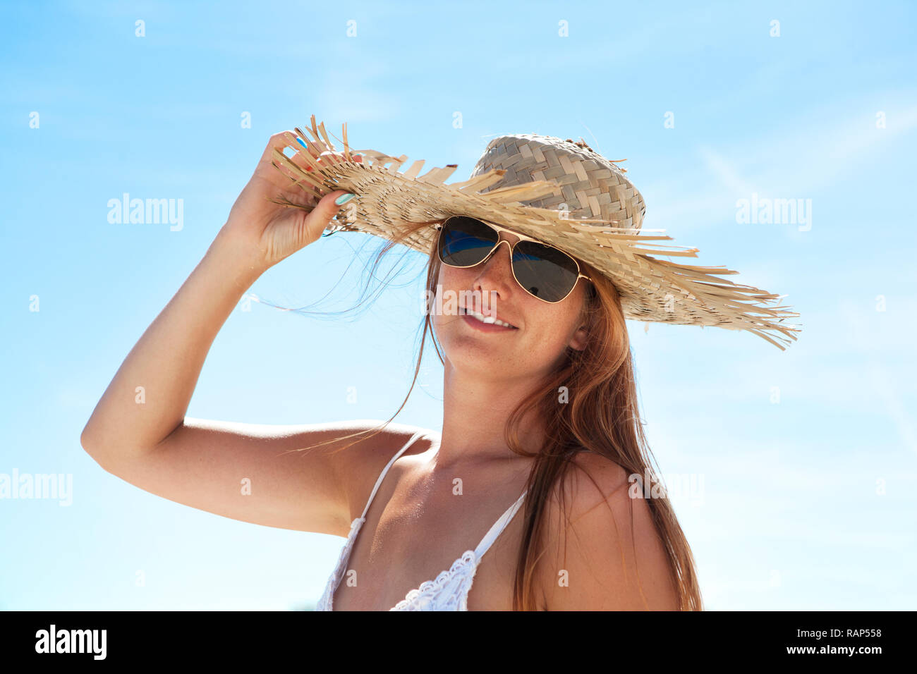 a young woman with long brown hair and sunglasses on the beach wearing a straw hat Stock Photo