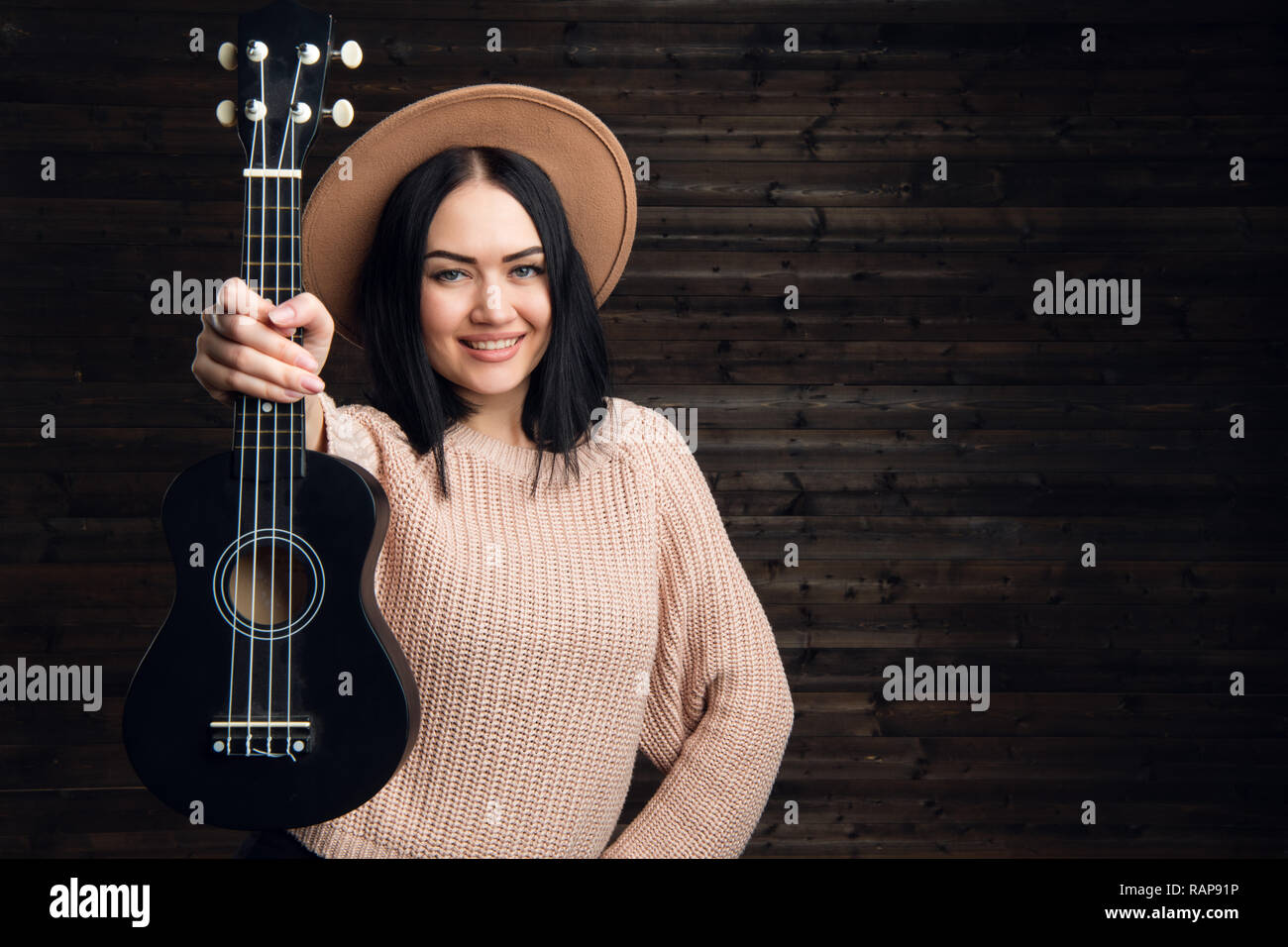 Young funny hipster girl having fun and playing on small ukulele guitar,  singing and dancing. wearing vintage hat, joy, positive mood. Dark wooden  wall background Stock Photo - Alamy