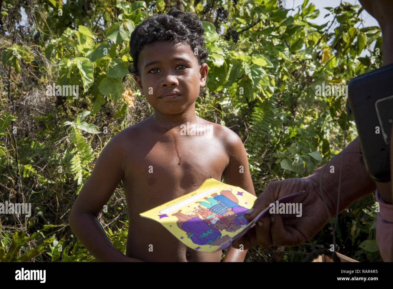 A father hands his child a book from a Low-Cost Low-Altitude bundle of humanitarian supplies during Operation Christmas Drop, Dec. 10, 2018, on Fais Island, Federated States of Micronesia. Although Fais has a small airstrip, the islanders rarely get visitors, mail, or supplies, and look towards OCD each year for the resupply of clothing, fishing, educational, and medical supplies. Stock Photo