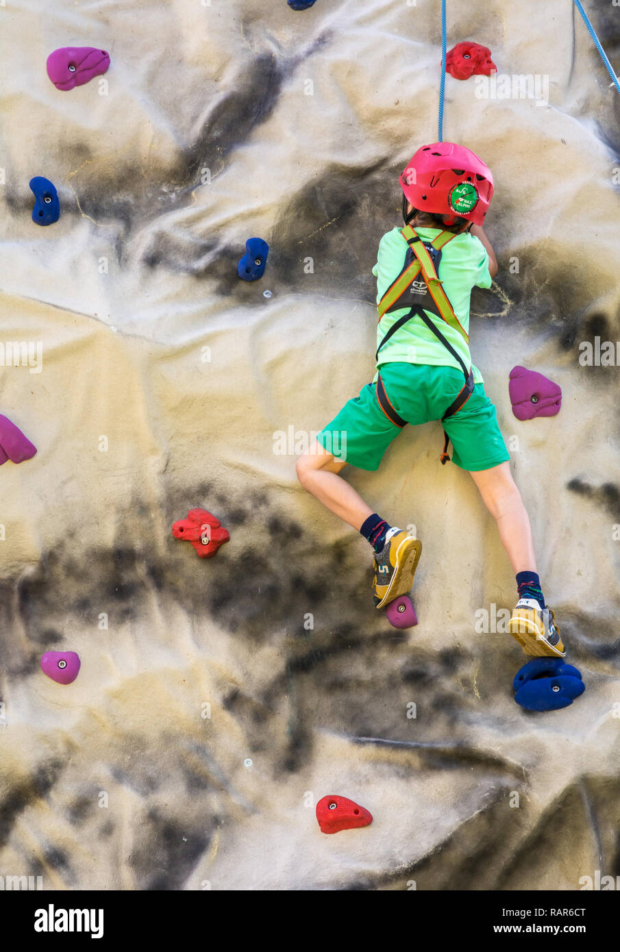 shot of little boy in a harness climbing a wall with grips. Stock Photo