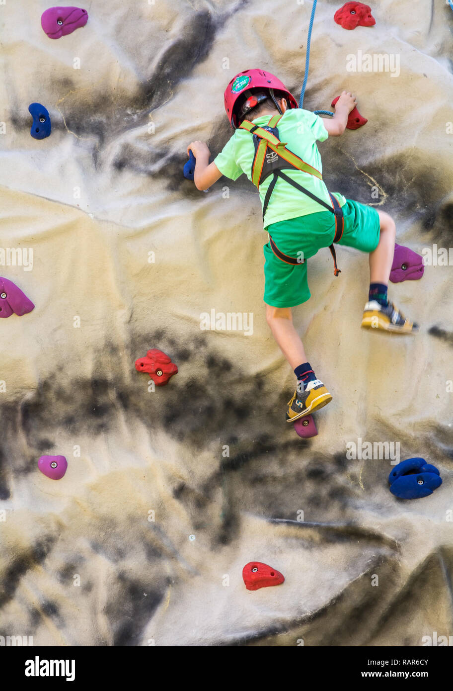 shot of little boy in a harness climbing a wall with grips. Stock Photo
