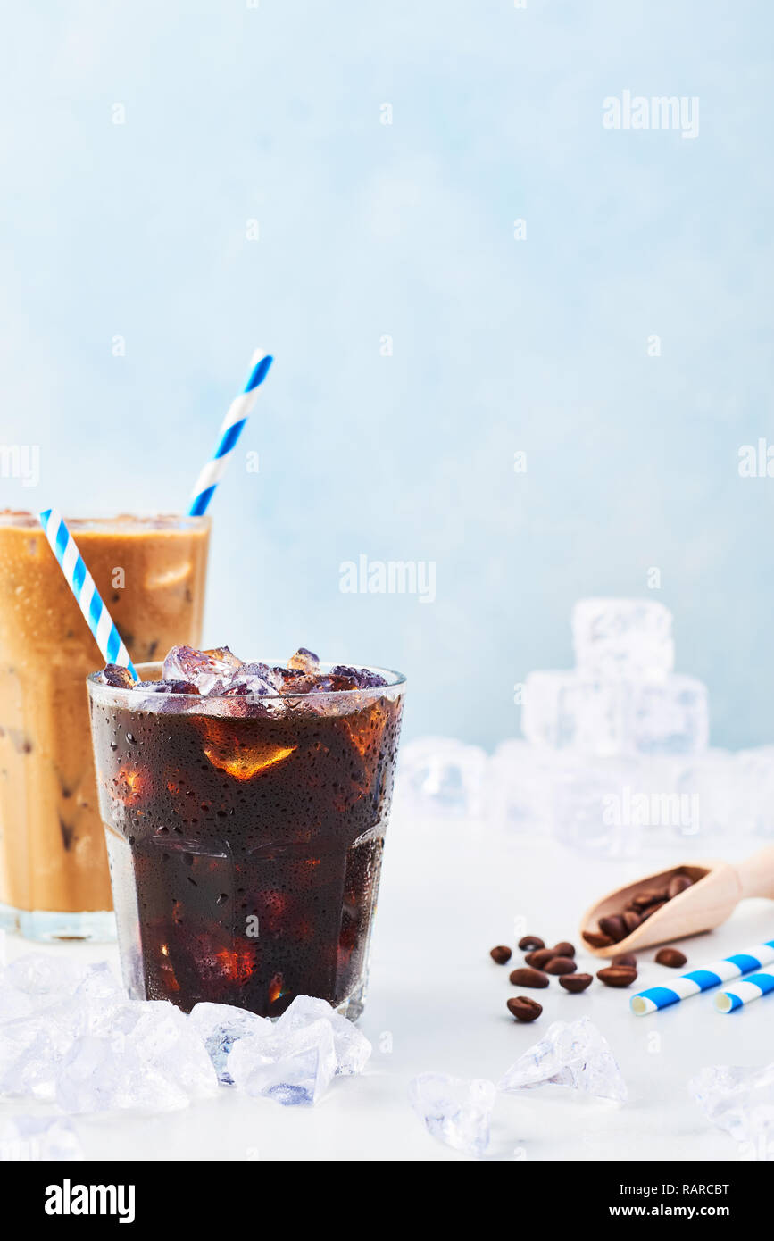 Summer drink iced coffee in a glass and ice coffee with cream in a tall glass, coffee beans and straws surrounded by ice on white marble table over bl Stock Photo
