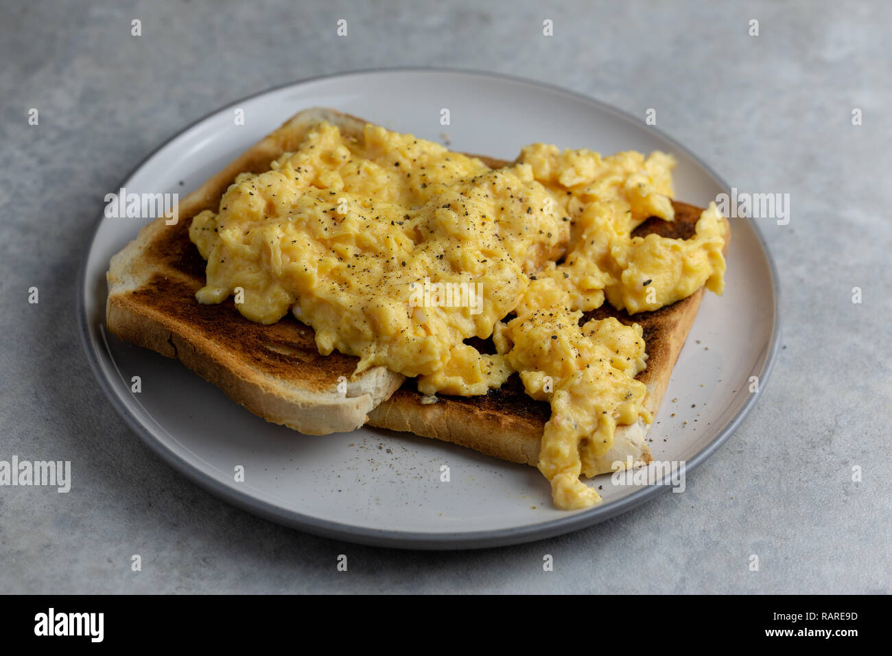 Scrambled eggs with black pepper on two pieces of toast on a white plate with a grey rim on a simple grey and white marbled background Stock Photo