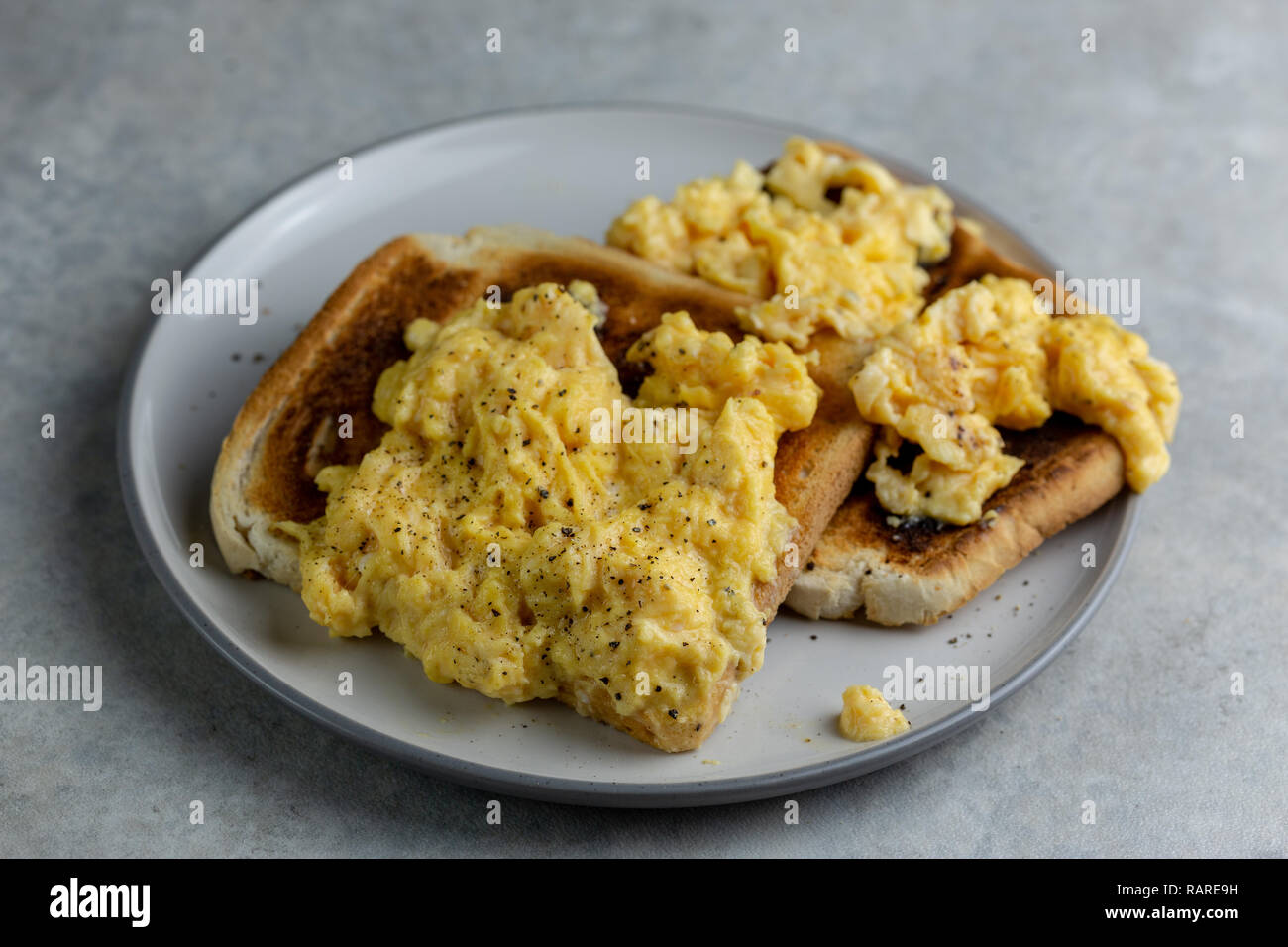 Scrambled eggs with black pepper on two pieces of toast on a white plate with a grey rim on a simple grey and white marbled background Stock Photo