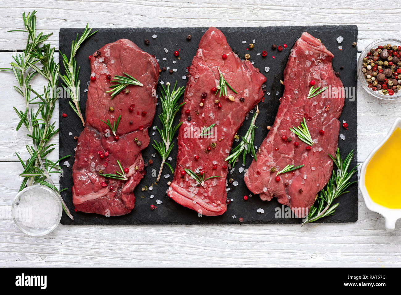 Raw meat, beef steak with spices, olive oil and rosemary on black slate cutting board over wooden background. top view Stock Photo