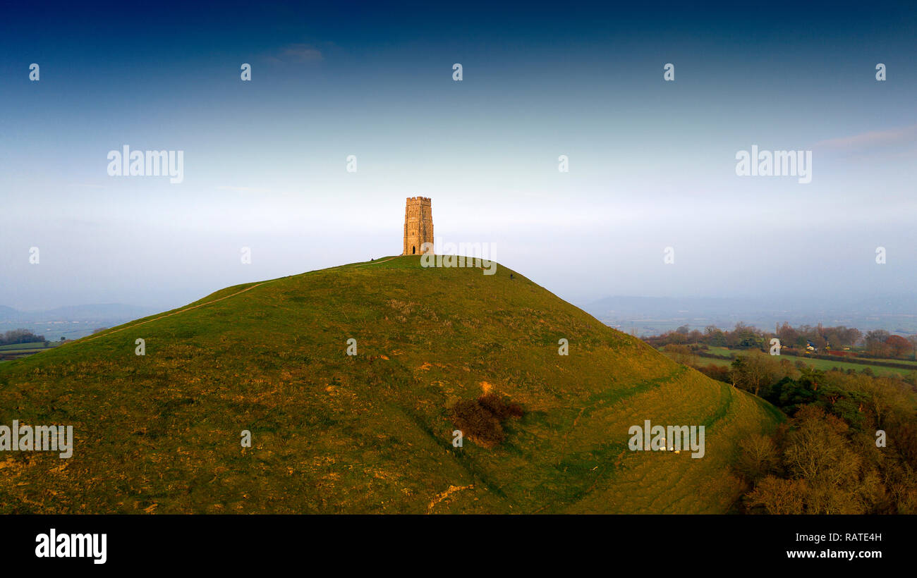 Aerial view of  Glastonbury Tor in the English county of Somerset, topped by the roofless St Michael's Tower, a Grade I listed building. The whole site is managed by the National Trust, and has been designated a scheduled monument. The conical hill of clay and Blue Lias rises from the Somerset Levels. The Tor is mentioned in Celtic Mythology, particularly in myths linked to King Arthur, and has a number of other enduring mythological and spiritual associations Stock Photo