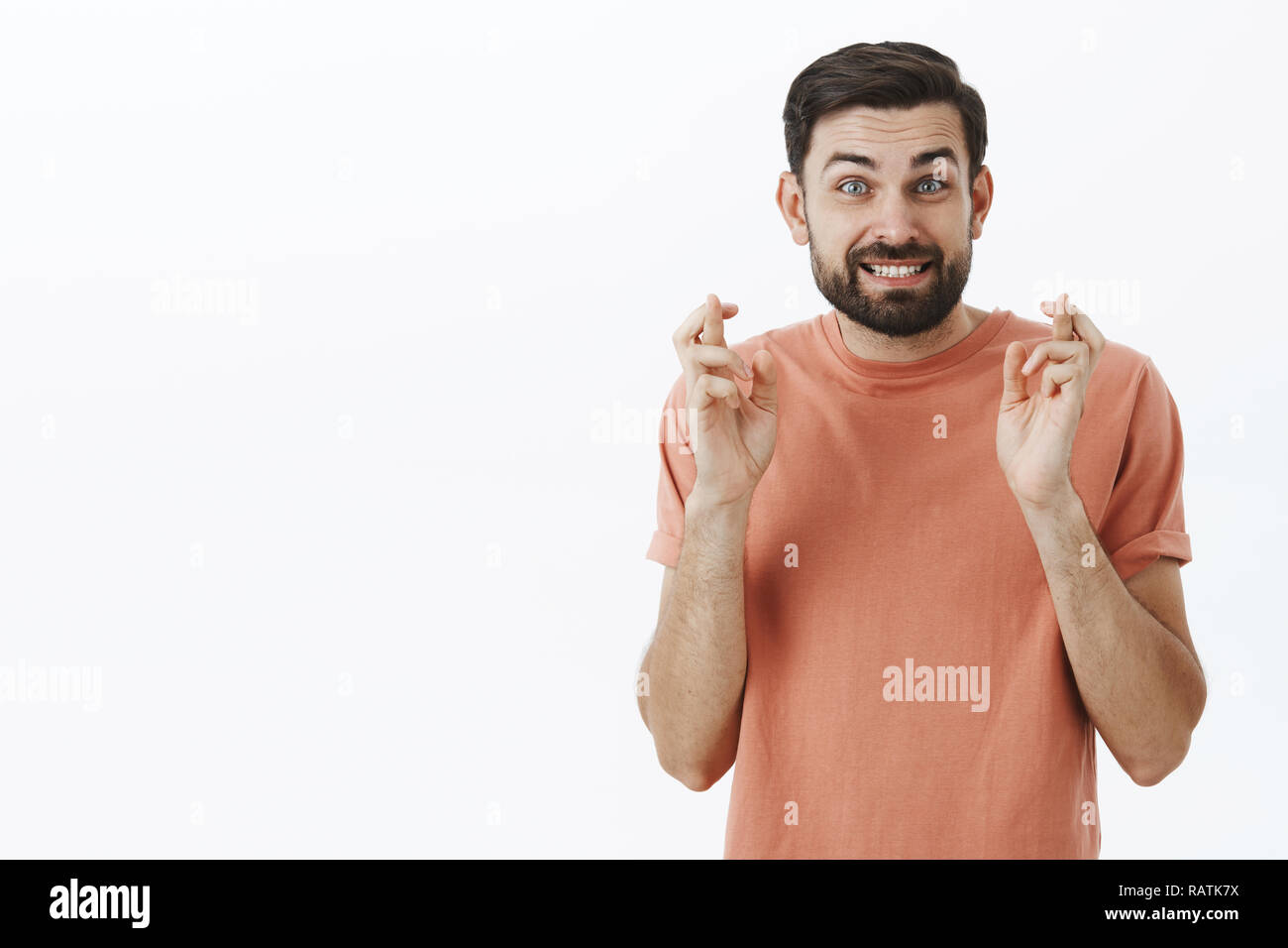 Studio shot of hopeful optimistic cute boyfriend with beard stooping and smiling with worry crossing fingers for good luck making wish for desired dream come true praying against white background Stock Photo