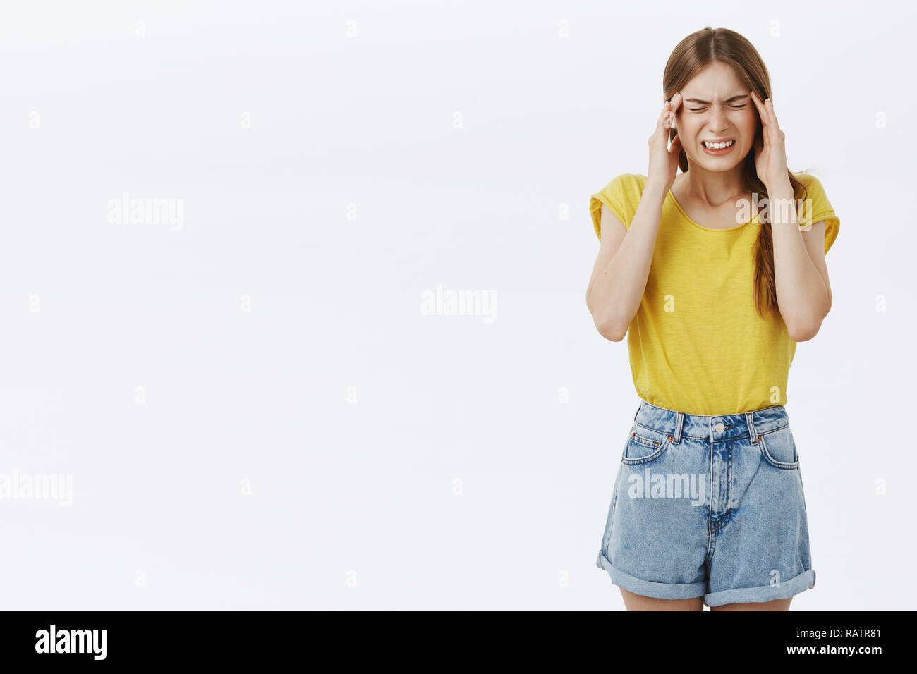 Intense good-looking caucasian slim woman in yellow t-shirt and shorts clenching teeth closing eyes and frowning holding hands on temples feeling unfocused and troubled raising tension while thinking Stock Photo