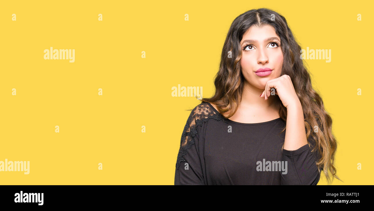 Beautiful young woman with long hair with hand on chin thinking about question, pensive expression. Smiling with thoughtful face. Doubt concept. Stock Photo