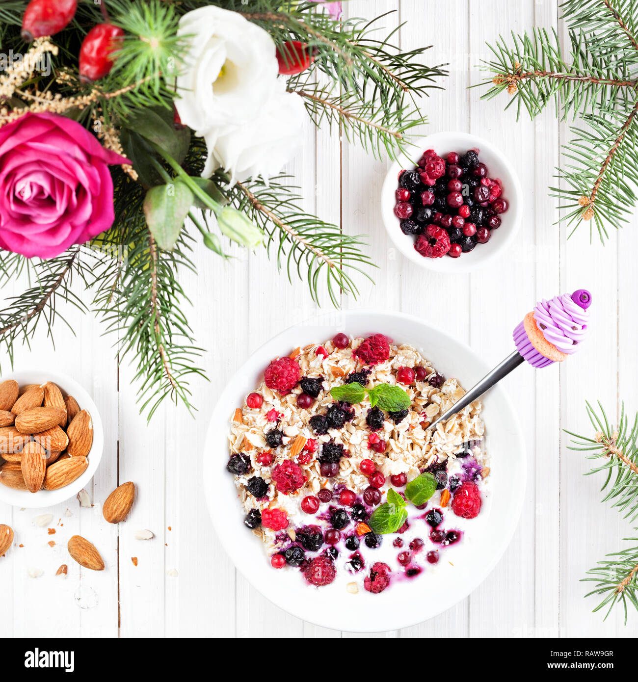 Decorative spoon with violet cupcake in the bowl with oats, berries and almond near winter flower bouquet on wooden white background top view Stock Photo