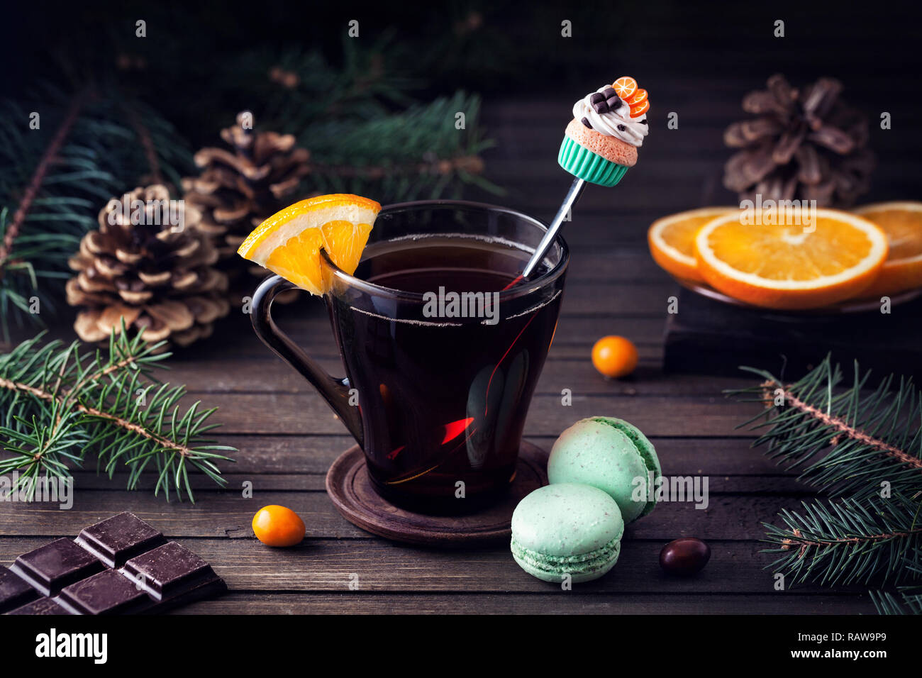 Decorative spoon with cupcake in the glass with tea and oranges slice near green macarons and chocolate on wooden background at Christmas time Stock Photo