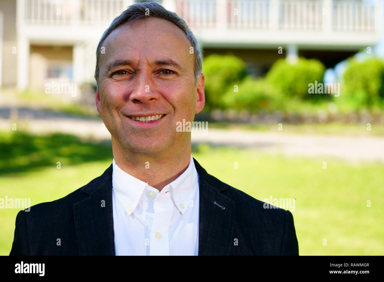 An outdoor close up portrait of a proud caucasian man wearing a white shirt, a black coat and smiling at the camera. Stock Photo