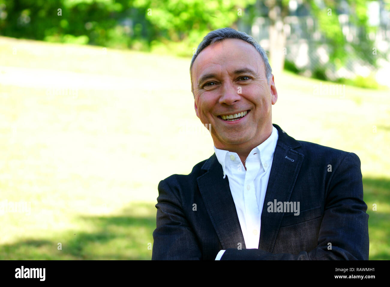 An outdoor close up portrait of a proud caucasian man wearing a white shirt, a black coat and smiling at the camera. Stock Photo