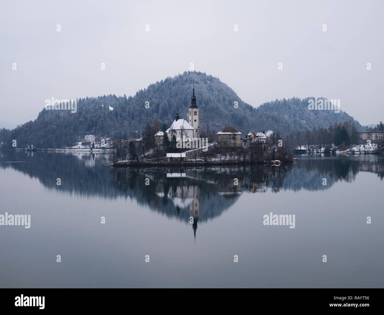 Winter grey moody Bled lake shot, with reflection of Pilgrimage Church of the Assumption of Maria covered in snow with almost no colour cast Stock Photo