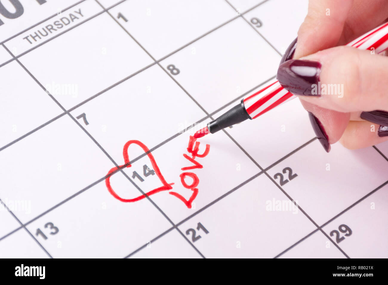 Girl hand with red felt pen drawing heart shape and writing word Love in calendar. Love and Valentines day holiday concept. Close up Stock Photo
