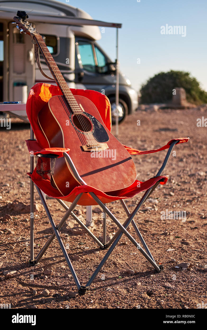 Vertical view red acoustic guitar on a folding chair near recreational vehicle motor home trailer, no people. Hobby traveling and lifestyle concept Stock Photo
