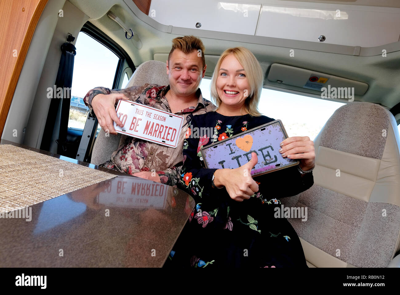 Cheerful middle age married positive travelers couple sitting inside of camper holds showing hippy retro styled number plate Stock Photo