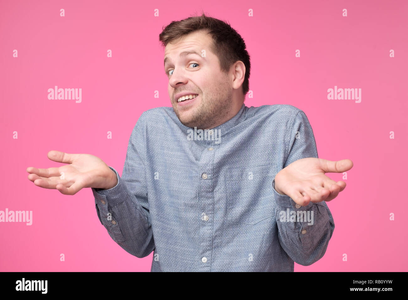 Uncertain young stylish man wears blue shirt, shrugs shoulders being puzzled or confused. Caucasian unsure male make gestures doubtfully with hands st Stock Photo