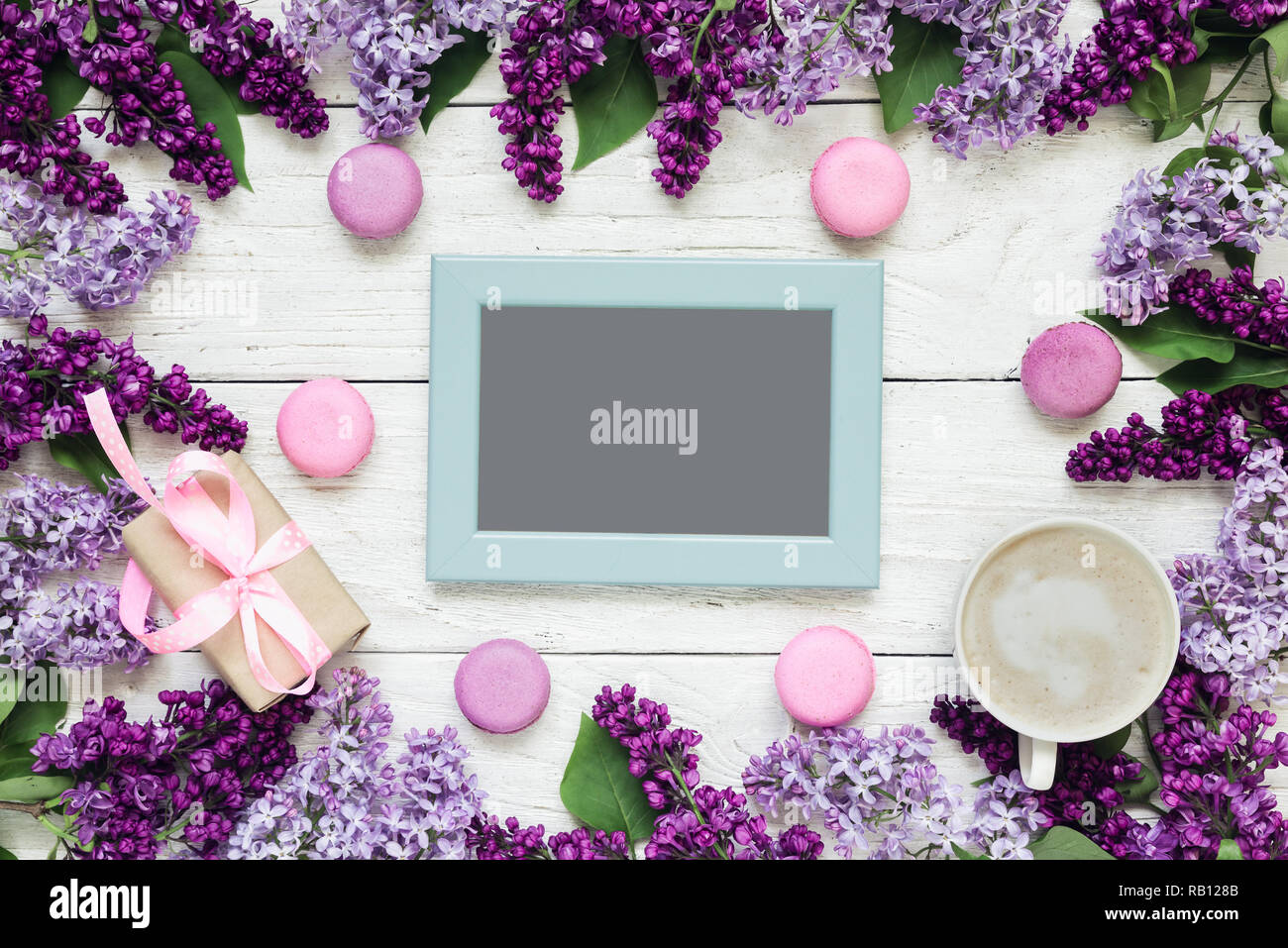 blank photo card in frame made of lilac flowers with coffee cup, gift box and macaroons over white wooden table. mock up. flat lay. top view. mothers  Stock Photo
